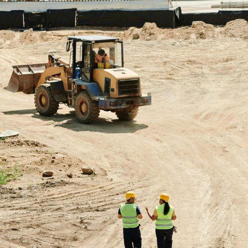 Construction workers in safety gear walking at a busy site with a backhoe in operation.
